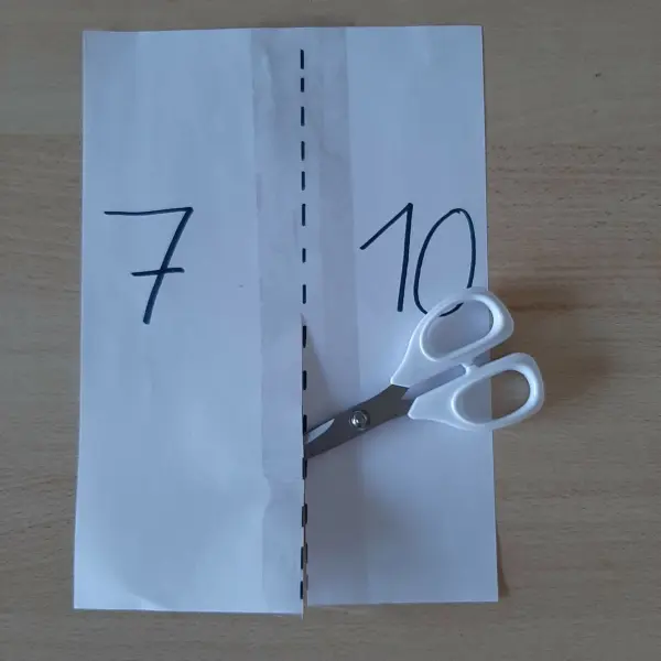 Ein Blatt Papier mit den Zahlen 7 und 10 wird mit einer Schere entlang einer Linie geschnitten; es liegt auf einem Tisch.
