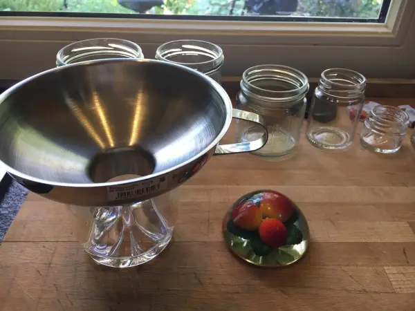 Ein silberner Trichter steht auf einem Glassockel, umgeben von leeren Einmachgläsern auf einem Holzbrett.
