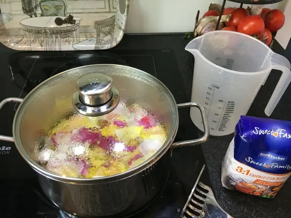 Ein Topf mit Duftrosen-Gelee kocht auf dem Herd, während ein Messbecher und Zucker daneben stehen.