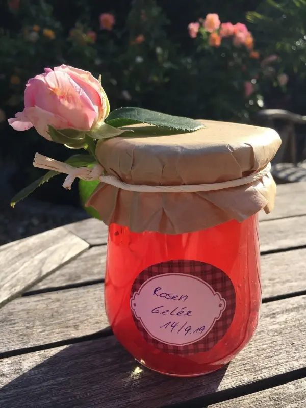 Ein Glas mit rosafarbenem Duftrosen-Gelee steht auf einem Holztisch, dekoriert mit einer Rose und einem Etikett.