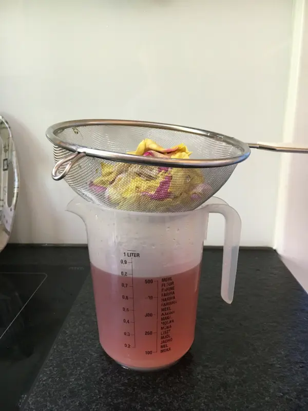 Ein Messbecher steht auf einer dunklen Oberfläche, während Duftrosen-Gelee durch ein Sieb gefiltert wird.