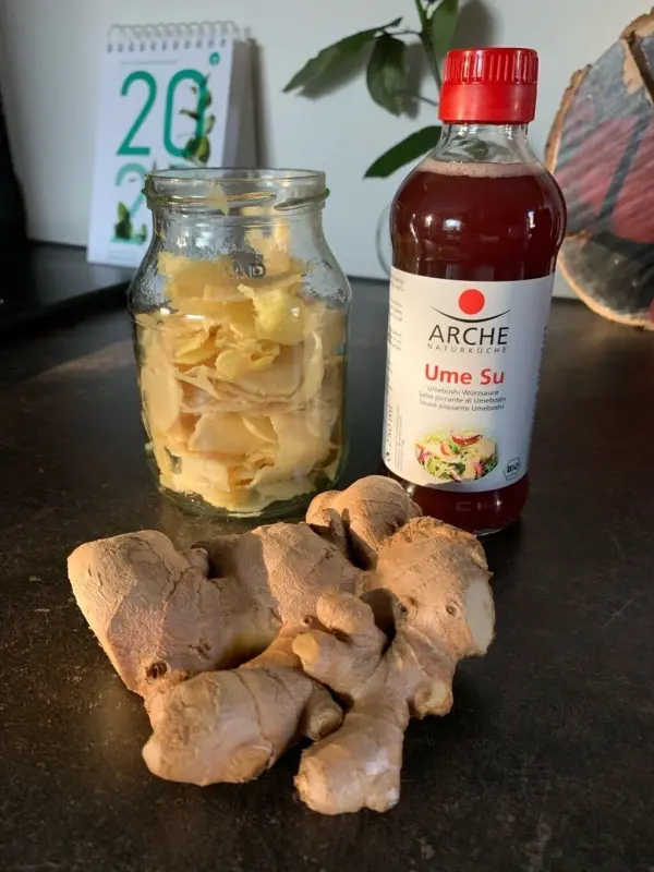 Frischer Ingwer liegt neben einem Glas mit eingelegtem Ingwer und einer Flasche milder, salziger Fruchtsoße auf einer dunklen Oberfläche.