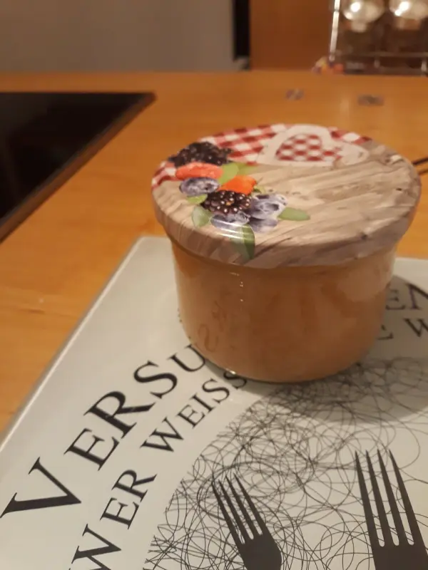 Ein Glas mit dekorativem Holzdeckel steht auf einem Tisch, umgeben von einer modernen Küchenoberfläche, wahrscheinlich gefüllt mit Bananen-Birnen-Marmelade.