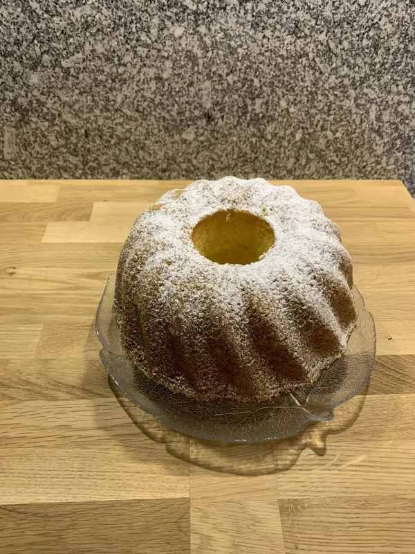 Ein Quark-Gugelhupf steht auf einem Glassockel, bestäubt mit Puderzucker, vor einer rustikalen Holztischoberfläche.