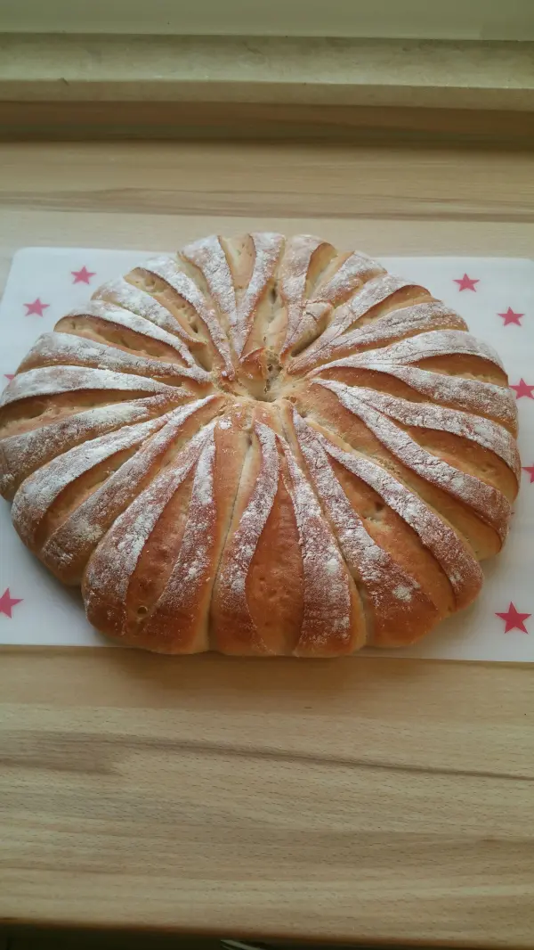 Ein runder Brötchen-Kranz liegt auf einem Holzbrett, leicht mit Puderzucker bestäubt, in einer hellen Küche.