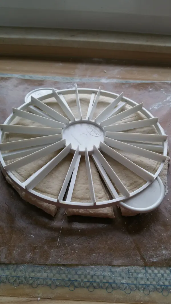 Ein runder Brötchen-Kranz aus Teig liegt auf einer bemehlten Arbeitsfläche und wird mit einem speziellen Cutter in Stücke geschnitten.