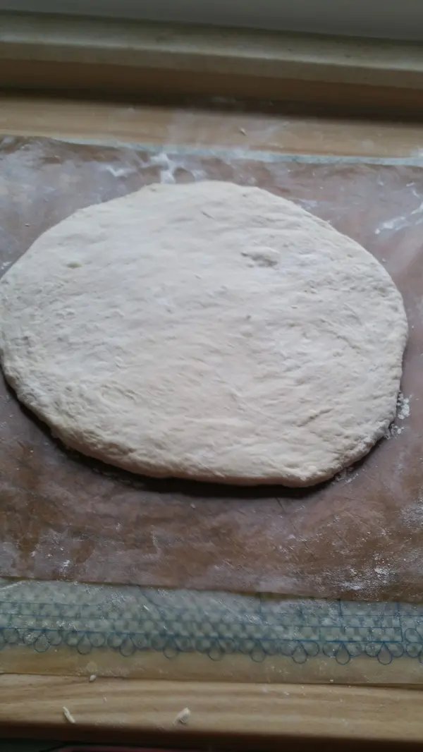 Ein runder, ausgerollter Brötchen-Teig liegt auf einer bemehlten Arbeitsfläche, bereit zum Weiterverarbeiten.