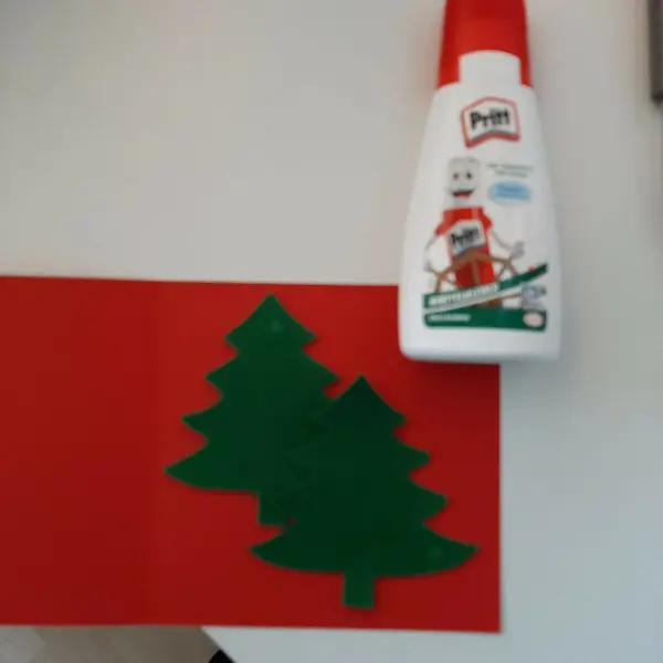 Eine rote Karte liegt auf dem Tisch, während grüne Weihnachtsbäume und weiße Kleberflasche zur Dekoration bereitstehen.
