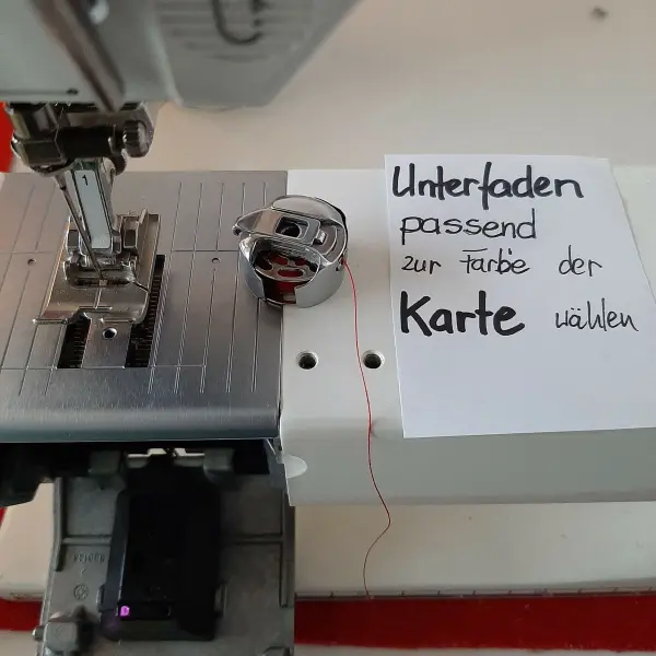 Eine Nähmaschine zeigt eine Anleitung auf einem Zettel, der den passenden Unterfaden zur Farbe der Wunschkarte beschreibt.