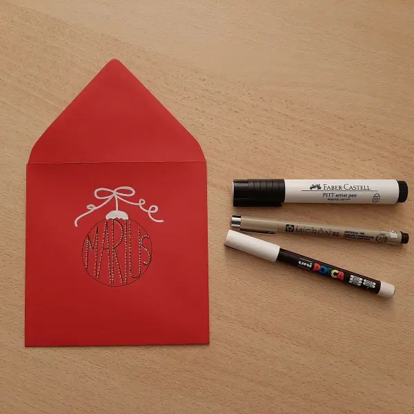 Eine rote Weihnachtskarte liegt auf einem Holzbrett, umgeben von drei Markern, bereit, eine Wunsch-Erfüller-Karte zu gestalten.