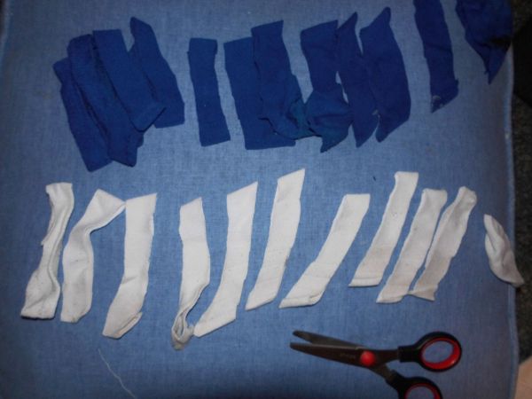 Bunte, zugeschnittene Stücke aus alten T-Shirts liegen auf einem Tisch, bereit zur Herstellung von Putzlappen. Schere daneben.