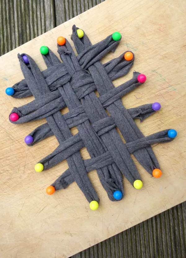 Ein graues T-Shirt ist zu einem Putzlappen gewebt, farbige Pins halten die Struktur auf einer Holzoberfläche fest.