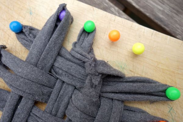 Graue Stoffstreifen sind zu einem Putzlappen geflochten, während bunte Pins als Befestigung dienen, auf einer Holzoberfläche.