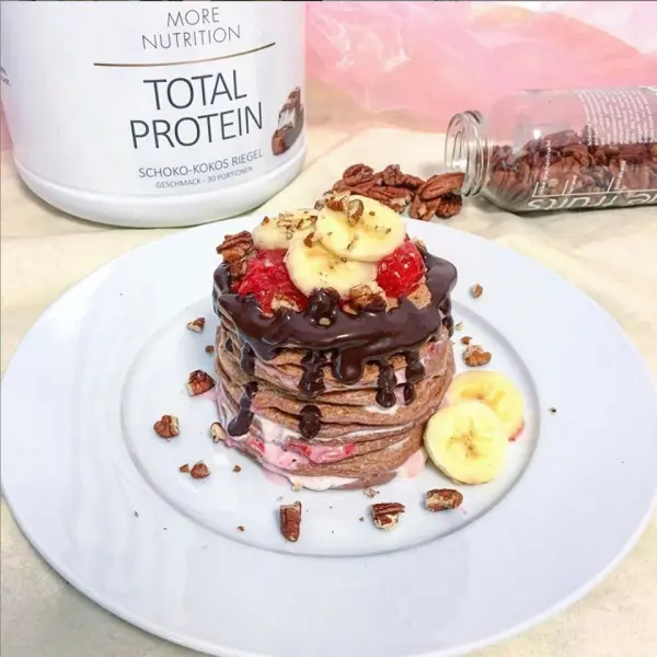 Pancakes liegen auf einem Teller und werden mit Schokolade, Banane und Nüssen garniert, umgeben von Proteinpulver.