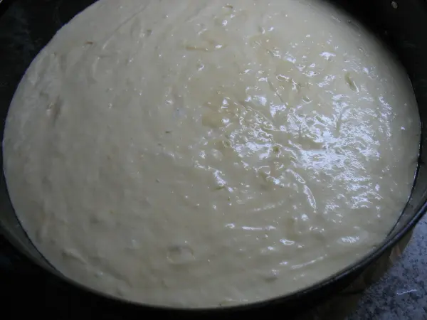 Eine helle, glatte Zitronentorte-Teigmasse ruht in einer runden Backform auf einer dunklen Arbeitsfläche, bereit zum Backen.