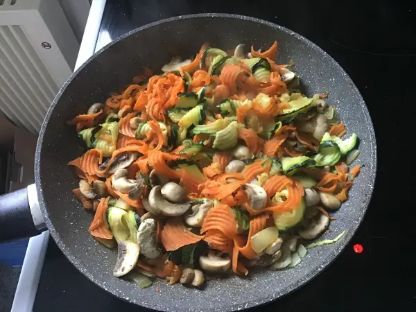 Bunte Gemüse-Nudeln braten in einer Pfanne, umgeben von frisch geschnittenem Gemüse wie Karotten, Zucchini und Pilzen.