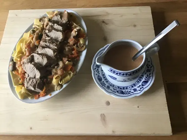 Auf einem Holzbrett liegt ein Gericht mit Schweinefilet auf Gemüsenudeln, dazu eine kleine Käsesoße in einer weißen Schüssel.