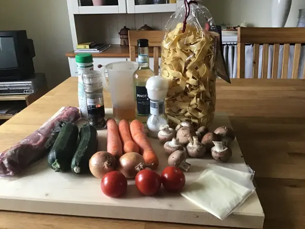Ein Schweinefilet liegt auf einem Holzbrett, umgeben von Gemüse wie Zucchini, Karotten, Tomaten und Pilzen, sowie Pasta.
