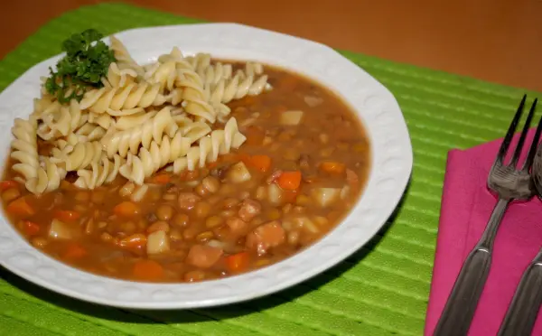 Nudeln liegen dekorativ neben einer schmackhaften Linsensuppe auf einem grünen Tischset, umgeben von Besteck.
