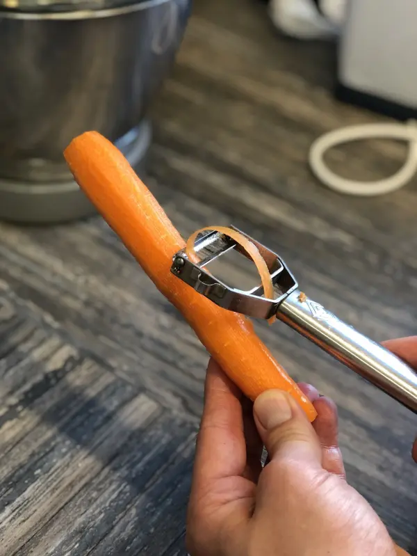 Eine Hand schält eine orange Karotte mit einem Schäler auf einer Holzoberfläche neben einer Küchenmaschine.