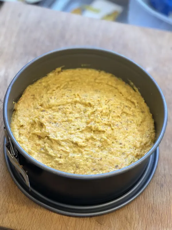 Ein Karottenkuchen in einer runden Backform steht auf einem Holzbrett, bereit zum Backen in einer modernen Küche.