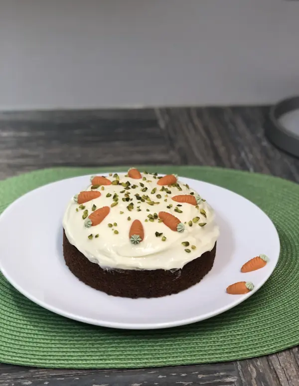 Ein saftiger Karottenkuchen steht auf einem weißen Teller, dekoriert mit Karotten und Pistazien, auf einem grünen Platz.