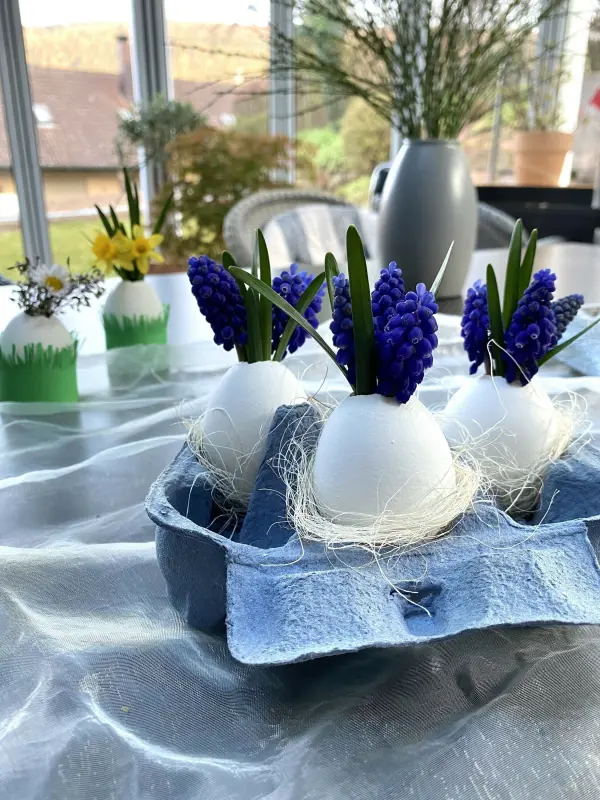Eierväschen mit blauen Blumen stehen in einem Eierkarton auf einem Tisch, umgeben von frischen Frühlingsdekorationen.