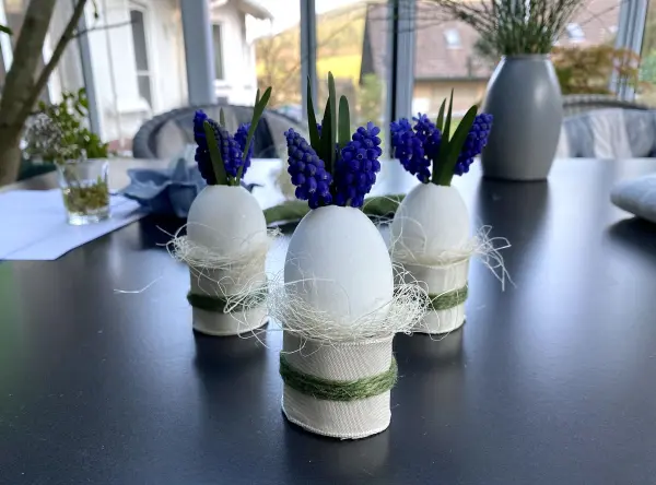 Weiße Eierväschen mit kleinen blauen Blumen stehen auf einem Tisch, umgeben von Sonnenschein und einem modernen Raum.