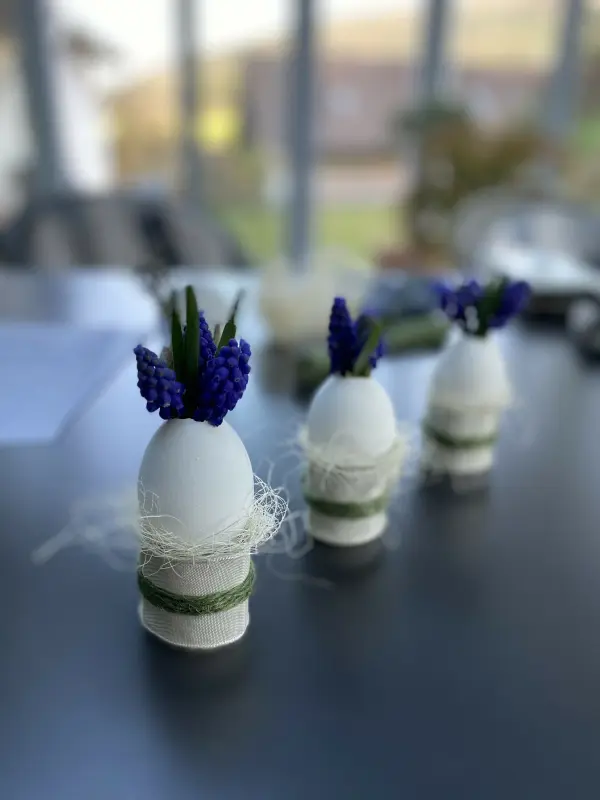 Drei dekorierte Eierväschen stehen auf einem Tisch, geschmückt mit lila Blumen und umgeben von einer hellen, modernen Umgebung.