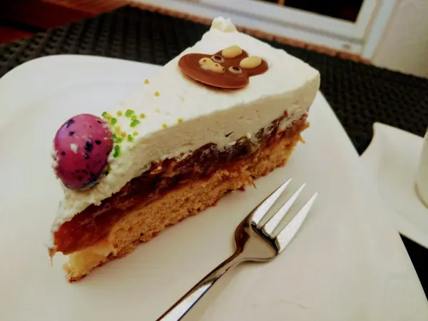 Ein Stück Rhabarberkuchen liegt auf einem Teller, dekoriert mit Sahne und bunten Plättchen, auf einem Tisch.
