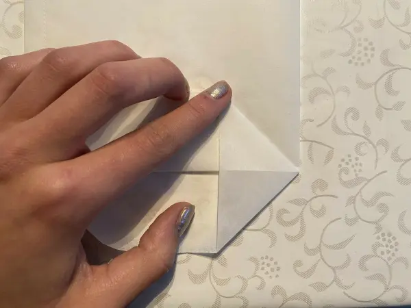 Eine Hand hält einen weißen Bogen, während sie eine Ecke zu einer Lichttüte für den Muttertag faltet, auf einem gemusterten Untergrund.