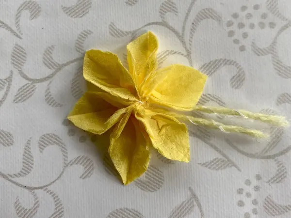Eine gelbe Papierblume liegt auf einem hellen, gemusterten Hintergrund und sorgt für eine fröhliche DIY-Deko-Idee zum Muttertag.