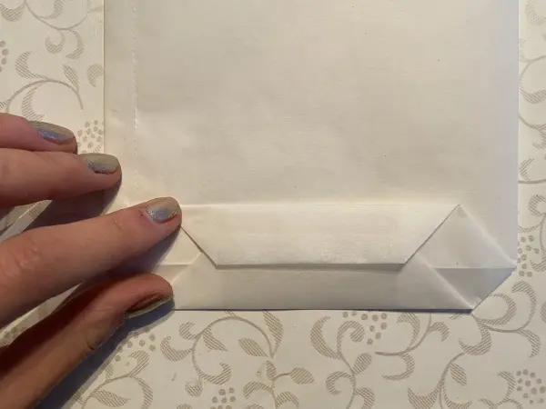 Eine Hand faltet die untere Ecke einer weißen Tüte, um eine DIY-Lichttüte für den Muttertag zu gestalten.