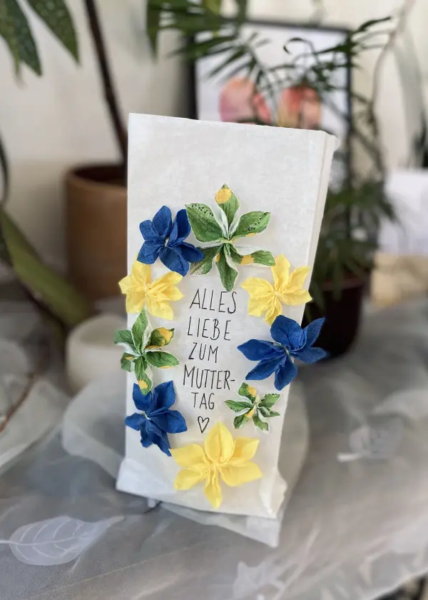 Eine dekorative Lichttüte ziert mit bunten Papierblüten den Schriftzug „ALLES LIEBE ZUM MUTTERTAG“ im Raum.