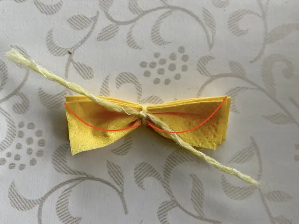 Ein gelbes, gefaltetes Papierstück in Form einer Schleife ist mit einer Schnur in der Mitte zusammengebunden und auf einem hellen, gemusterten Untergrund platziert.