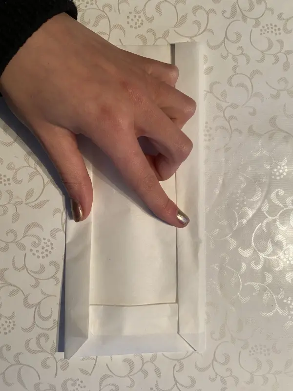 Eine Hand faltet eine Papierlichttüte auf einem eleganten Tisch mit floralem Design, ideal für DIY zum Muttertag.