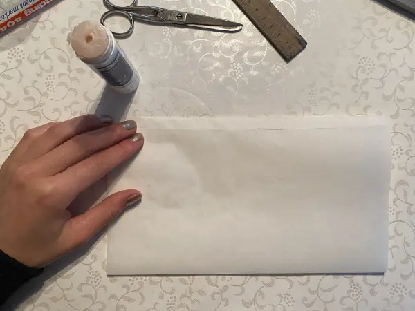 Eine Hand hält eine weiße Tüte auf einem Tisch, während eine Schere und ein Kleber daneben liegen. Ideal für DIY zum Muttertag.