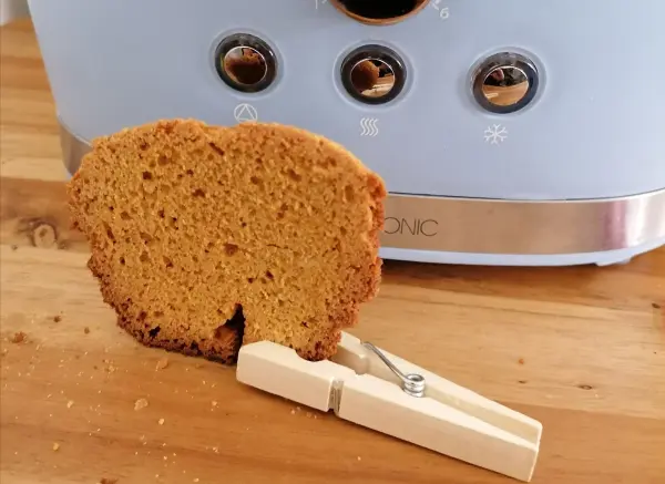 Eine knusprige Toastscheibe steht mit einer Wäscheklammer an einem Toaster auf einer Holzoberfläche.
