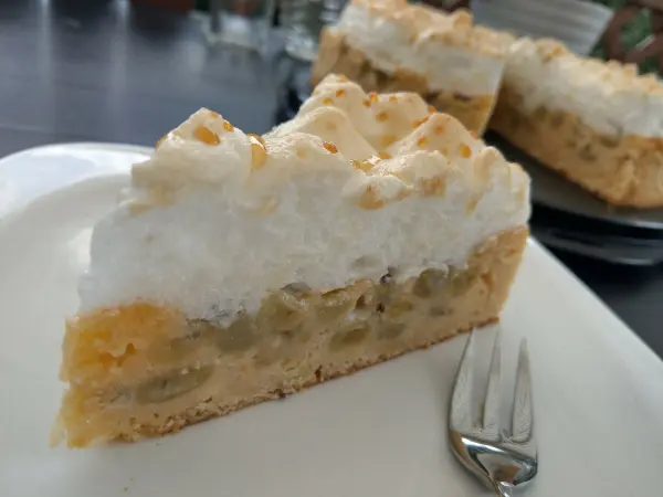 Ein Stück Stachelbeerkuchen mit Baiser liegt auf einem Teller, während im Hintergrund weitere Kuchenstücke sichtbar sind.