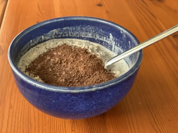Eine blaue Schüssel mit Müsli steht auf einem Holztisch, während ein Löffel im Müsli steckt, das mit Schokoladenpulver bestreut ist.