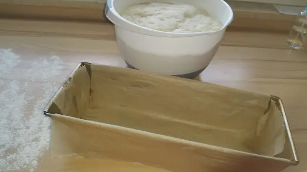 Eine Backform steht leer auf einer Holzoberfläche, während ein Schüssel mit Teig im Hintergrund auf das Toastbrot warten.