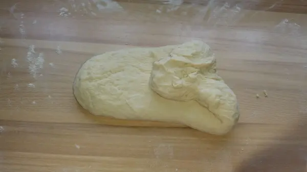 Ein rohes Stück Brot-Teig liegt auf einer holzfarbenen Arbeitsfläche, umgeben von etwas Mehl für das Backen von Toastbrot.