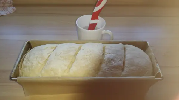Frisches Toastbrot liegt in einer Kastenform auf einem Tisch, daneben steht eine Tasse mit einem Strohhalm.