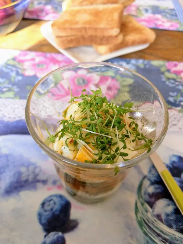 Ein Glas mit Eiern und frischer Kresse steht auf einem bunten Tisch mit Blumenmuster, daneben Toastscheiben.