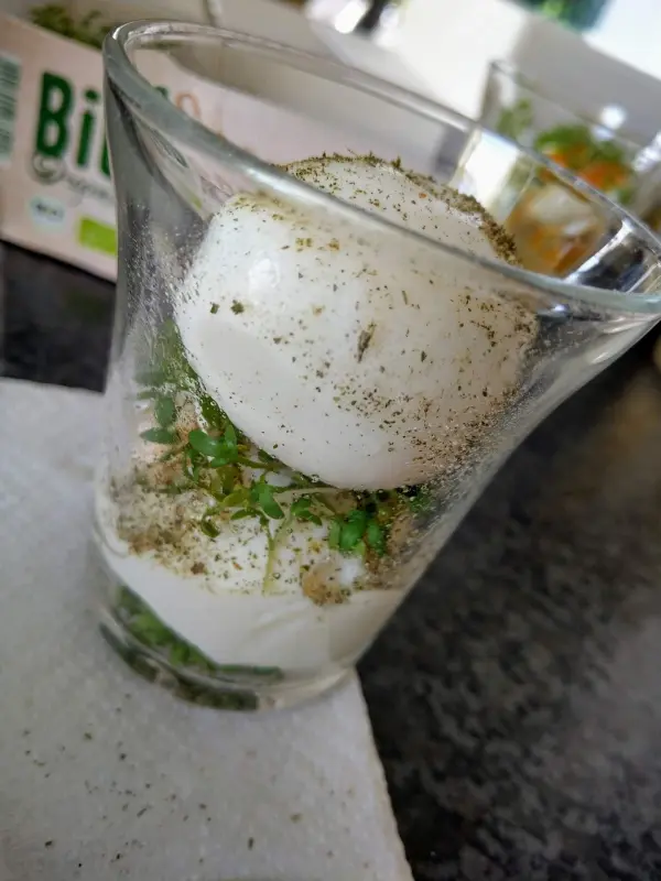 Ein Glas enthält ein gekochtes Ei, umgeben von feiner Kresse und Gewürzen. Es steht auf einem Tisch.