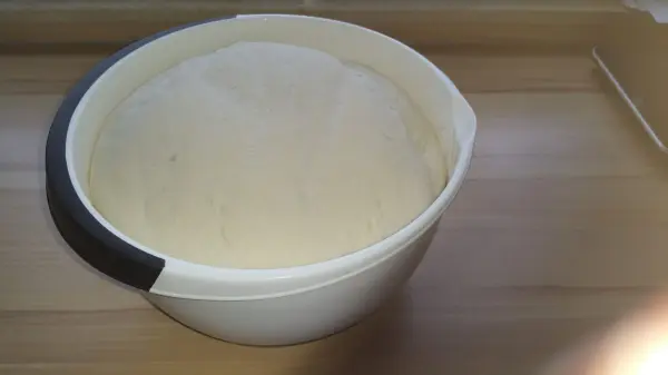 Eine Schüssel mit Hefe-Teig steht auf einem Holzbrett. Der Teig ist aufgegangen und bereit zum weiteren Verarbeiten für die Weizenbrötchen.