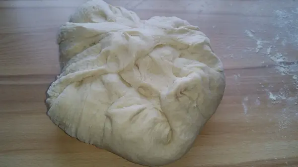 Ein weiches Brotstück liegt auf einem Holzbrett, bearbeitet und bereit für das Formen von knusprigen Weizenbrötchen.