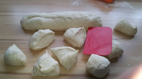 Teigstücke liegen auf einer Holzoberfläche, ein länglicher Teigstrang und ein pinker Teigschaber sind sichtbar.