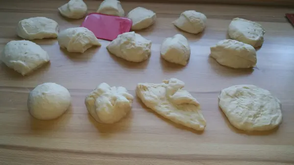 Eine Gruppe roher Weizenbrötchen liegt auf einer Holzplatte, bereit zum Backen, mit einem bunten Teigschaber daneben.