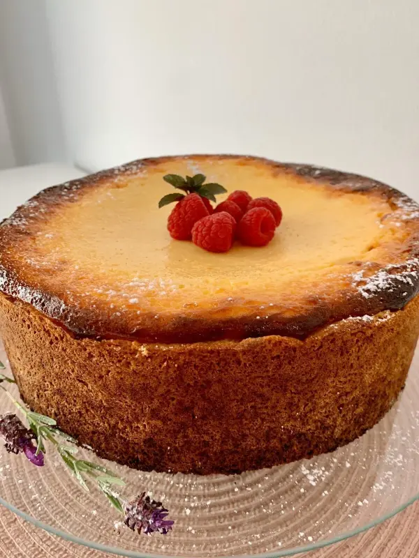 Ein saftiger Käsekuchen steht auf einer Glasplatte, dekoriert mit frischen Himbeeren und einem grünen Blatt, in einem hellen Raum.